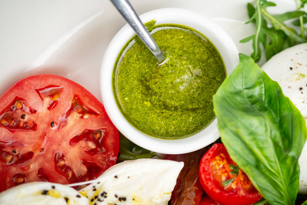 Basil pesto sauce in a small bowl, with fresh basil leaves, tomato and mozzarella cheese, close up - Photo, Image