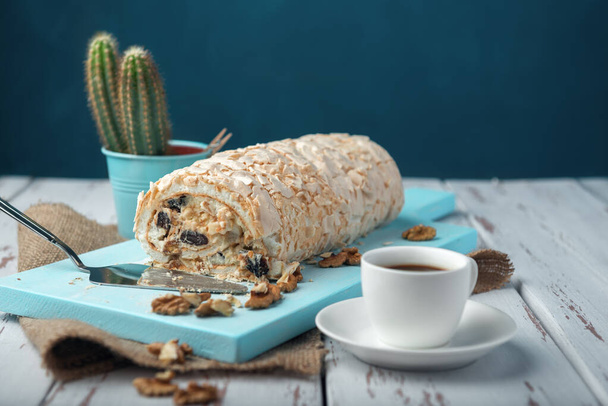 A cup of coffee meringue roll on a white vintage wooden kitchen table with burlap napkin. Meringue pie decorated with prunes and walnuts on blue cutting board. - Photo, Image