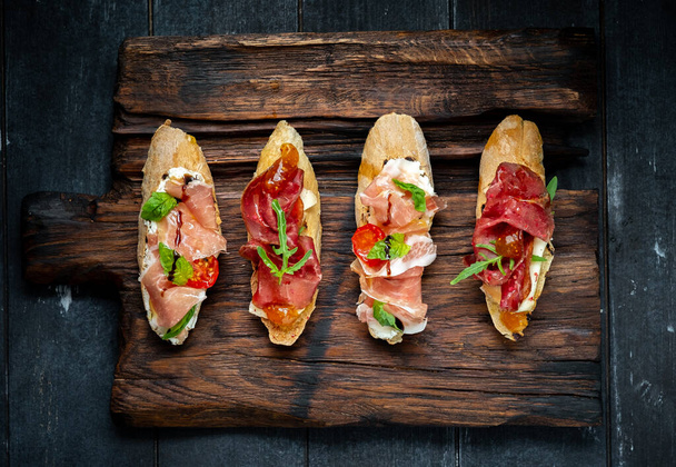 Traditional Bruschetta with Parma dried ham and prosciutto. Italian antipasti set sandwiches with jamon, cottage cheese, arugula, and sun-dried tomatoes on a rustic wooden Board Top view - Fotó, kép