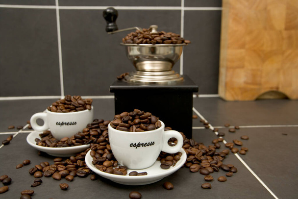 Natureza morta de grãos de café torrados, xícaras de café expresso e moedor em uma superfície de trabalho de cozinha em azulejos
 - Foto, Imagem