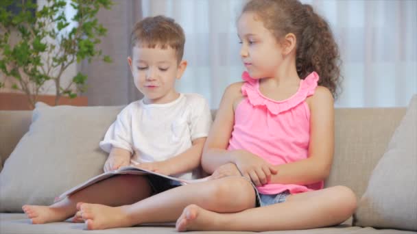 Cute children,sister with a younger brother, looking at a book magazine textbook dictionary tablet read fairy tales, kids are reading books sitting on the couch,preschool and school education. - Materiaali, video