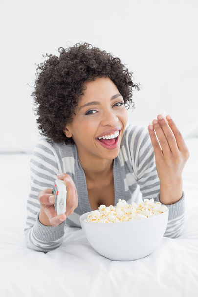 Cheerful woman with remote control and popcorn bowl in bed - Foto, imagen