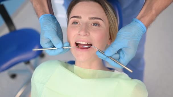 Diş hekimi muayenehanesinde bir hastanın dişlerini diş malzemesi kaşığıyla muayene ediyor. Stomatoloji ve sağlık hizmetleri kavramı. Tek kullanımlık tıbbi maskeli doktor, gülümseyen mutlu kadın.. - Video, Çekim