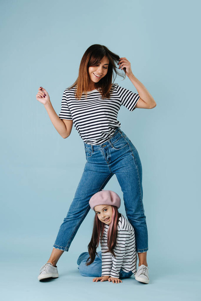 Two fashion lovers, mom and daughter wearing same clothes over blue background. They have jeans and black and white striped shirt on them. Mom in a flattering pose, looking down at her daughter. - Photo, image