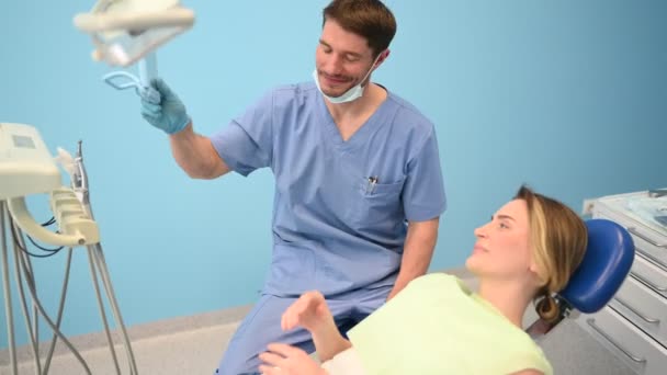 Der Zahnarzt zeigt dem Patienten die Ergebnisse der Behandlung mit einem Spiegel und untersucht die Zähne mit zahnärztlichen Geräten in der Zahnarztpraxis. Junge hübsche männliche Arzt in medizinische Gesichtsmaske und lächelnde glückliche Frau. - Filmmaterial, Video