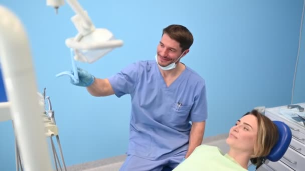 Der Zahnarzt zeigt dem Patienten die Ergebnisse der Behandlung mit einem Spiegel und untersucht die Zähne mit zahnärztlichen Geräten in der Zahnarztpraxis. Junge hübsche männliche Arzt in medizinische Gesichtsmaske und lächelnde glückliche Frau. - Filmmaterial, Video