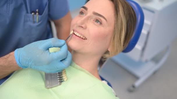 Οδοντίατρος ελέγχει το επίπεδο λεύκανσης των δοντιών του ασθενούς με το χρώμα ενός οδοντίατρου. Οδοντιατρικός εξοπλισμός στο οδοντιατρείο. Στοματολογία έννοια. Χέρια γιατρού σε ιατρικά γάντια και χαμογελαστή ευτυχισμένη γυναίκα. - Πλάνα, βίντεο
