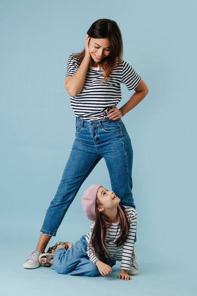 Two fashion lovers, mom and daughter wearing same clothes. They have jeans and black and white striped shirt on them. Mom in a flattering pose, looking down at her daughter, she looks back. - Φωτογραφία, εικόνα