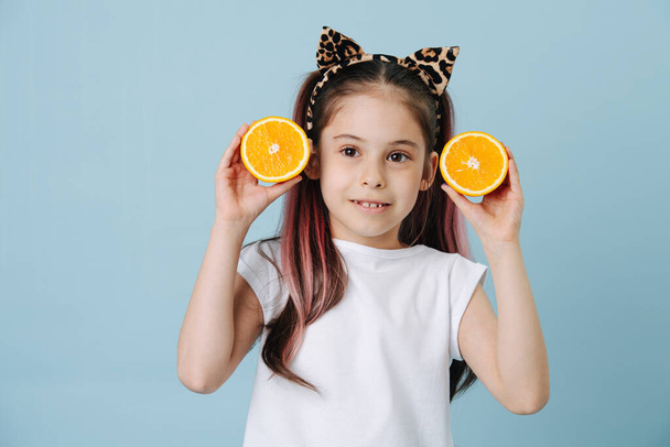 Portrait of a cute little girl with cat ears headband, posing for a photo in a studio. She's holding orange half-slices on each side of her face. Half length. - Foto, Imagen