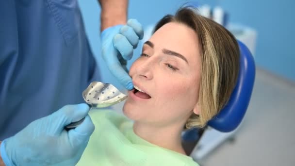 Стоматолог вивчає зуби пацієнта, використовуючи стоматологічне обладнання, вражаючу ложку в стоматологічному кабінеті. Стоматологія та концепція охорони здоров'я. Лікар в одноразовій медичній масці для обличчя, усміхнена щаслива жінка
. - Кадри, відео
