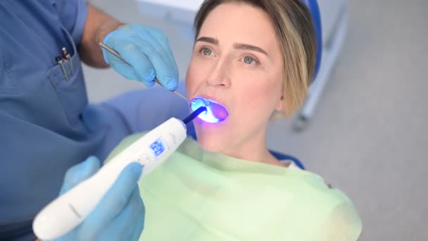 Οδοντίατρος που χρησιμοποιεί οδοντιατρικό θεραπευτικό ελαφρύ εξοπλισμό για την πλήρωση, αφού εξετάσει τα δόντια ενός ασθενούς στο οδοντιατρικό γραφείο. Στοματολογία και υγειονομική περίθαλψη έννοια. Κοντινό πλάνο νεαρή ευτυχισμένη γυναίκα στην οδοντιατρική καρέκλα - Πλάνα, βίντεο