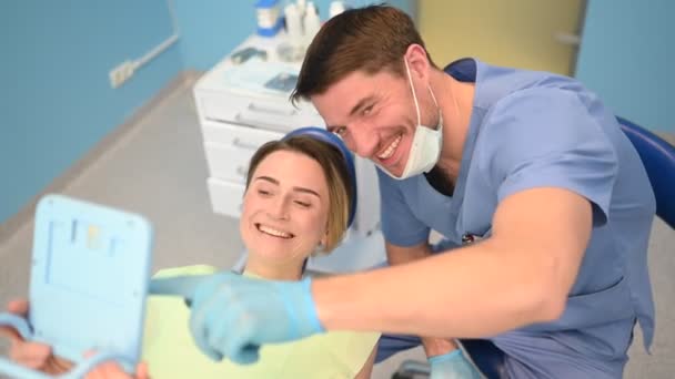 Tandarts toont de patiënt de resultaten van de behandeling met een spiegel, het onderzoeken van tanden met tandheelkundige apparatuur in tandheelkundige kantoor. Jonge knappe mannelijke arts in medisch gezichtsmasker en glimlachende gelukkige vrouw. - Video