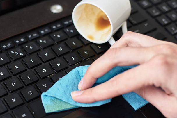Рука очищает пролитый кофе на клавиатуре ноутбука тряпкой
 - Фото, изображение