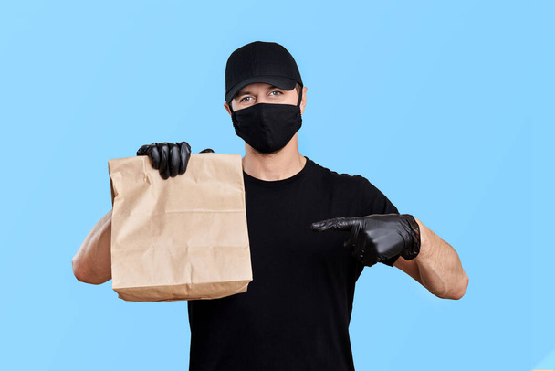 Concepto de entrega de alimentos. Mensajero en uniforme negro sostiene la bolsa de papel con comida para llevar sobre fondo azul
 - Foto, Imagen