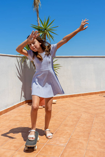 Pretty little girl in a striped dress playing with a skateboard on the terrace of her house. Fun concept - Photo, Image