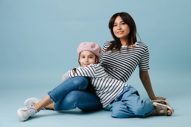 Two fashion lovers, mom and daughter wearing same clothes. They have jeans and black and white striped shirt on them. Mom sitting on the floor and leaning on the floor. Dau leaning on her. - Фото, изображение
