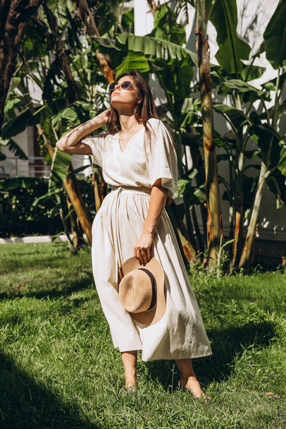 stylish young attractive woman dressed in a long linen dress with a hat photographed in the summer on a background of palm trees and the sea in the bright sun - Photo, Image