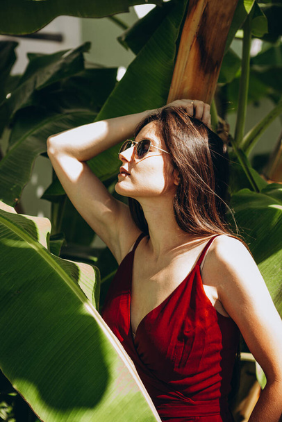 stylish young attractive woman dressed in a light sexy red dress photographed in the summer on a background of palm trees and the sea in the bright sun - Фото, изображение