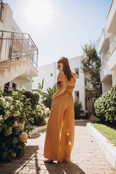 stylish young attractive woman dressed in an orange-colored trouser suit and sunglasses photographed in the summer on a background of palm trees and the sea in the bright sun - Foto, Imagem