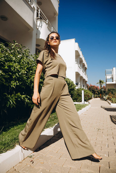 stylish young attractive woman dressed in dressed in a suit with a T-shirt and pants of olive color photographed in the summer on a background of palm trees and the sea in the bright sun - Φωτογραφία, εικόνα