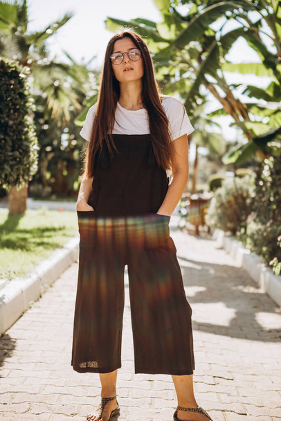 stylish young attractive woman dressed in in a stylish casual black jumpsuit and white T-shirt photographed in the summer on a background of palm trees and the sea in the bright sun - Foto, Bild