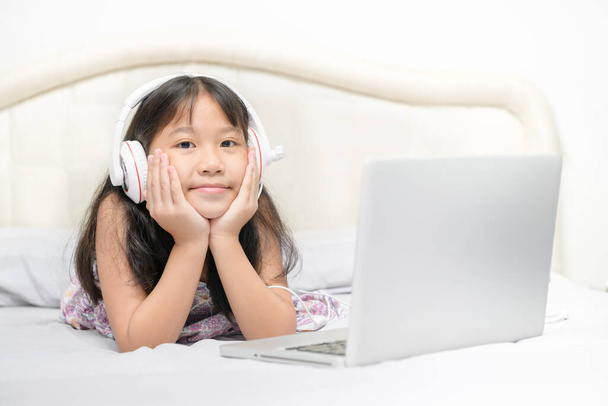 Asiático bonito menina usar fones de ouvido para aprender on-line usando um laptop no quarto. É uma Escola de Casa e Prevenção do Coronavírus (COVID-19). Educação conceitual
 - Foto, Imagem