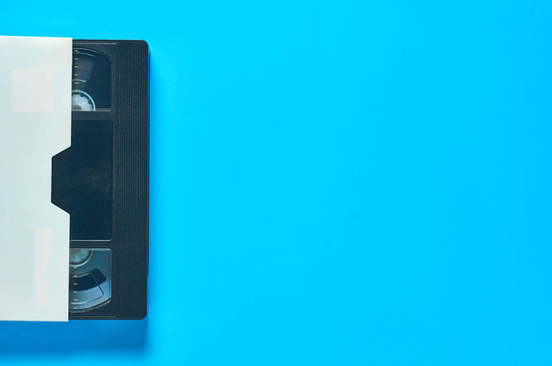 One black old vhs video cassette in white cardboard packaging on blue desk. Concept of 90s. Copy space. Top view - Фото, изображение