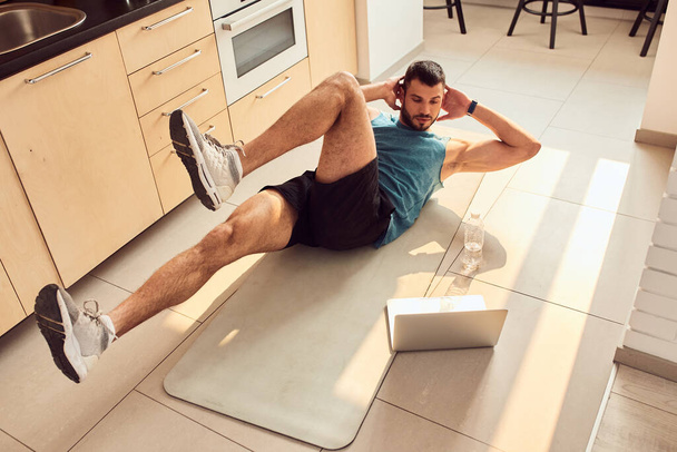 Handsome young man doing crunches and using laptop at home - Fotó, kép