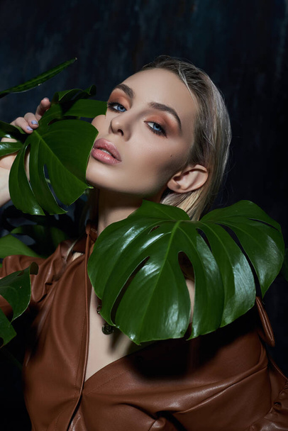 Portrait of a woman in tropical foliage. Wet hair, perfect figure and makeup. Beautiful blonde girl in leather dress and monstera leaves - Фото, зображення