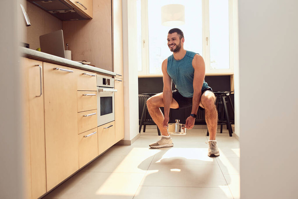 Joyful young man doing squats with cooking pot in kitchen - Foto, afbeelding