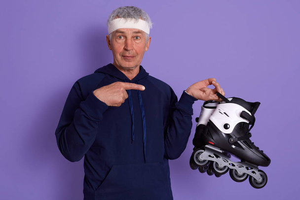 Closeup portrait of emotional charismatic old man making gesture, pointing with forefinger at roller skates, looking directly at camera, holding roller blades in hand. Active lifestyle concept. - Foto, afbeelding