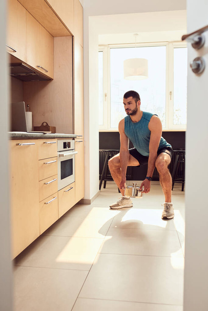 Handsome young man doing squats with cooking pot in kitchen - Foto, immagini