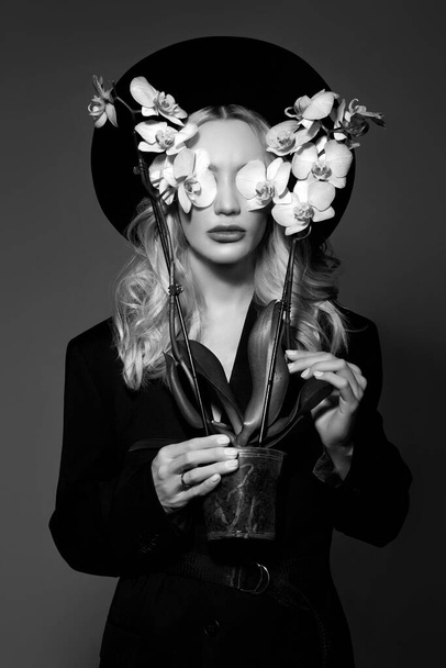 Portrait of a Blonde woman in a large round black hat, an Orchid flower in her hands - 写真・画像
