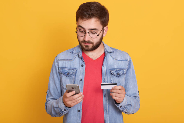 Bearded man paying with credit card on smart phone, looks concentrated, looking at device's screen, holding mobile phone and cardin hands, wearing denim jacket and red shirt, poses against yellow wall - Foto, afbeelding
