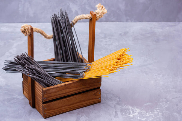 Black and yellow pasta in a wooden box. Donation wooden box with black spaghetti on a gray background. Spaghetti with cuttlefish ink. Food donation concept - Fotó, kép