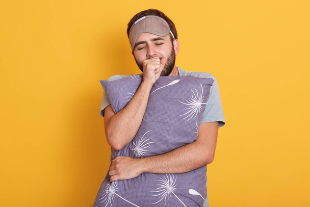 Young dark haired and bearded male yawning and covering mouth with his fist, keeps eyes closed, holding grey pillow in hands isolated over yellow background. - Foto, imagen