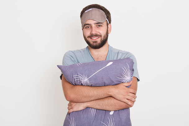 Closeup portrait of smiling bearded male embracing gray pillow, posing against white wall after waking up, bearded guy expressing positive emotions, looks happy. - Foto, Bild