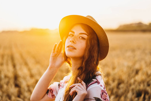 Portré egy gyönyörű fiatal nőről göndör hajjal és szeplős arccal. Nő ruha és kalap pózol búzamezőn naplementekor és a kamerába néz. - Fotó, kép