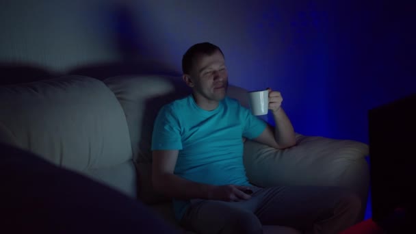 Ein Mann mit einem Becher heißem Tee sitzt spät am Abend vor dem Fernseher - Filmmaterial, Video