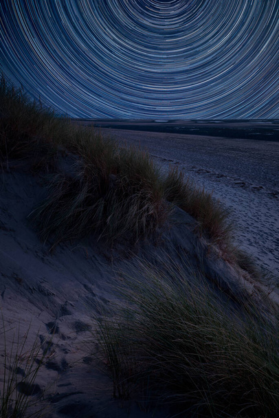 Imagen compuesta digital de senderos estelares alrededor de Polaris con paisaje de verano sobre dunas de arena cubiertas de hierba en la playa
 - Foto, Imagen