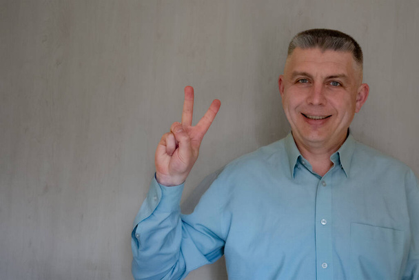 Middle aged man portrait. Victory sign gesture of 40 years man looking at camera and smiling. Blue business shirt is dressed. Happy face and positive emotions. Copy space background - Photo, Image