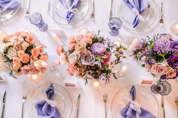 Presidium, mesa de banquete con un mantel blanco. Placas y cubiertos, vasos morados, servilletas, velas en candelabros de vidrio. Ramo de flores, astros, rosas, claveles, eustomas
. - Foto, imagen