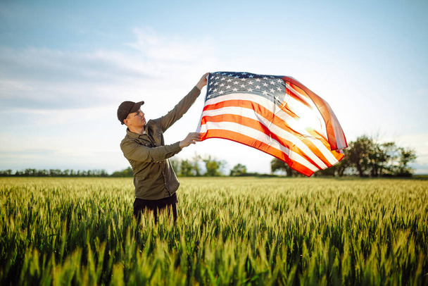 Young man wearing green shirt and cap lets the american flag fly on the wind at the green wheat field. Patriotic boy celebrates usa independence day on 4th of July with a national flag in his hands - Valokuva, kuva
