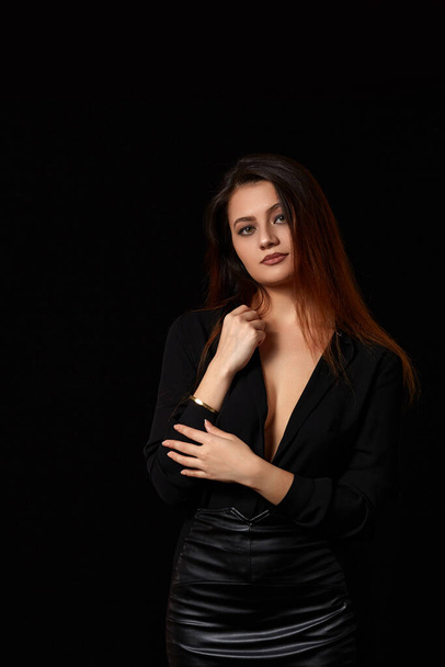 a young beautiful dark haired woman in a black unbuttoned shirt that revealed her Breasts and a black leather skirt on a black isolated background. - Fotó, kép