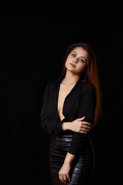 a young beautiful dark haired woman in a black unbuttoned shirt that revealed her Breasts and a black leather skirt on a black isolated background. - Fotoğraf, Görsel