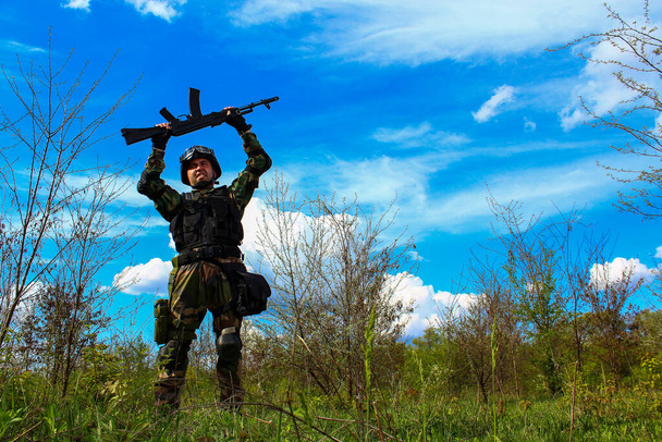 Een soldaat van de speciale troepen houdt een machinegeweer boven zijn hoofd en verheugt zich over de overwinning op een zeer mooie landschap achtergrond van de natuur met een blauwe lucht en groen - Foto, afbeelding