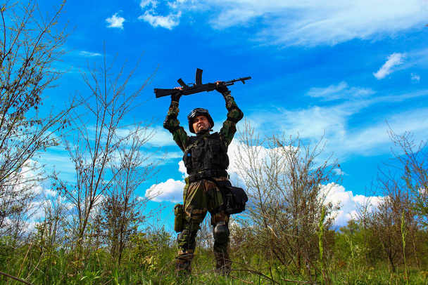 Un soldato delle forze speciali tiene una mitragliatrice sopra la testa gioisce della vittoria su uno sfondo paesaggistico molto bello della natura con un cielo blu e verdi
 - Foto, immagini