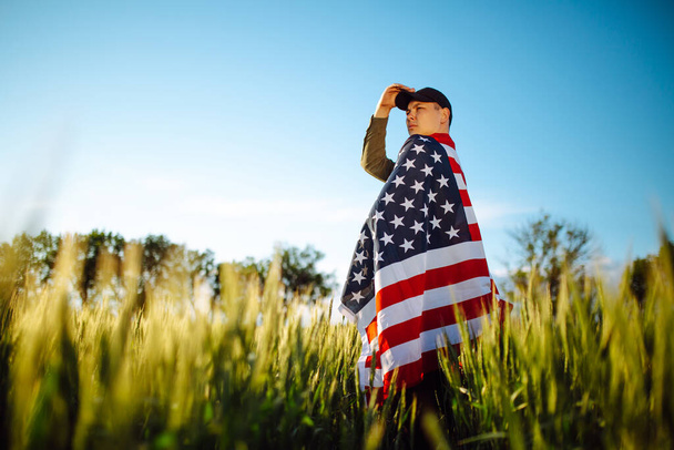 Egy zöld inget és sapkát viselő fiatalember áll az amerikai zászlóba csomagolva a zöld búzamezőn. A hazafias fiú július 4-én ünnepli a függetlenség napját egy nemzeti zászlóval a kezében. - Fotó, kép