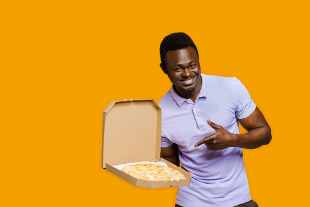 Finom pizzafutár az étteremből. Biztonságos szállítás. Pizza sajtos táblákkal reklámozza. Elszigetelt sárga háttér. A fiatal afrikai szakállas futár a pizzára mutat. - Fotó, kép