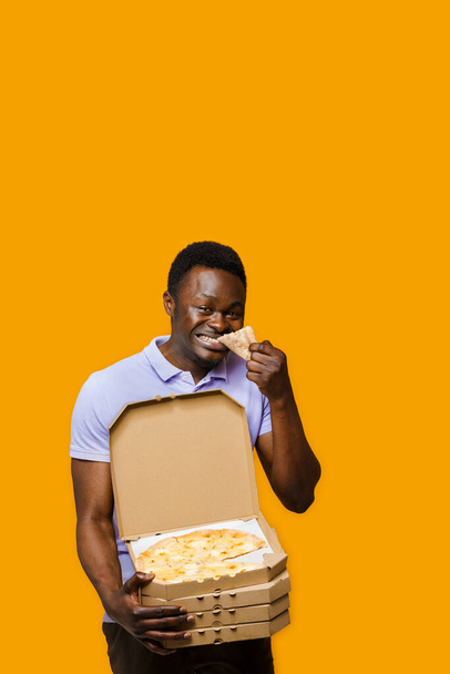 Funny black courier eats piece of pizza and holds 4 pizza boxes. 2+2 Big discount for customers. Tasty food pizza delivery from restaurant. Safety delivery. Pizza with cheese boards advert. - Photo, Image